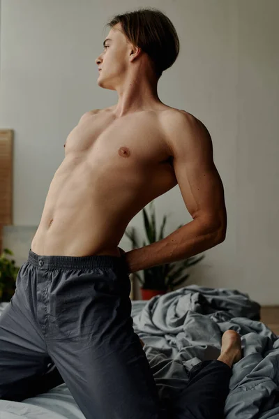 Sexy shirtless man in pajama pants stretching back while kneeling on bed — Stock Photo