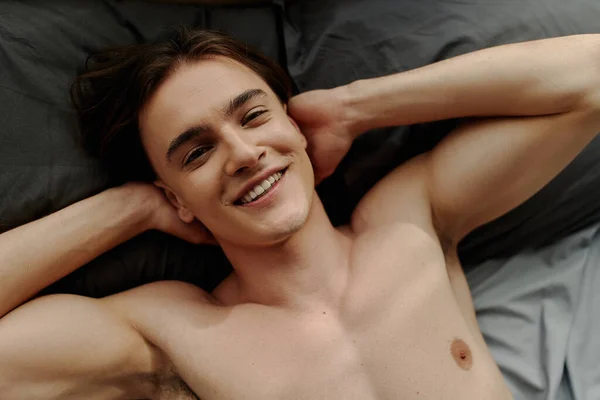 Vista superior del hombre acostado en la cama con las manos detrás de la cabeza y sonriendo a la cámara - foto de stock