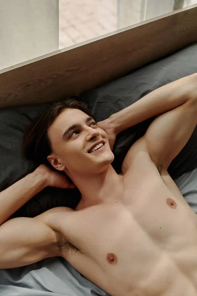 Top view of sexy young man looking away and smiling while lying on bed with hands behind head — Stock Photo