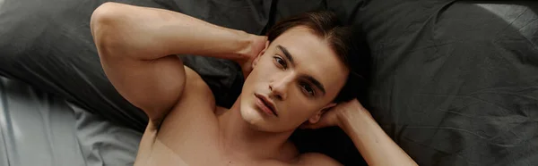 Vista superior del hombre sin camisa mirando a la cámara mientras está acostado en la cama con las manos detrás de la cabeza, pancarta - foto de stock