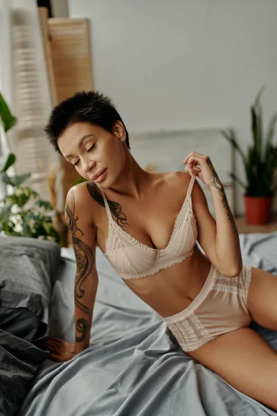 Femme tatouée passionnée en lingerie sexy assise sur le lit avec les yeux fermés — Photo de stock