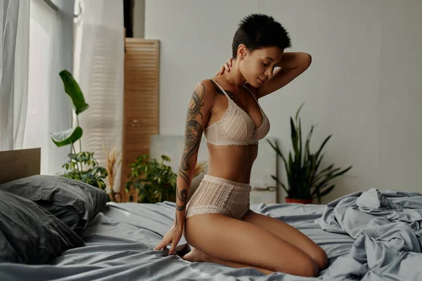 Side view of brunette woman in sexy underwear sitting with hand behind neck in bedroom — Stock Photo