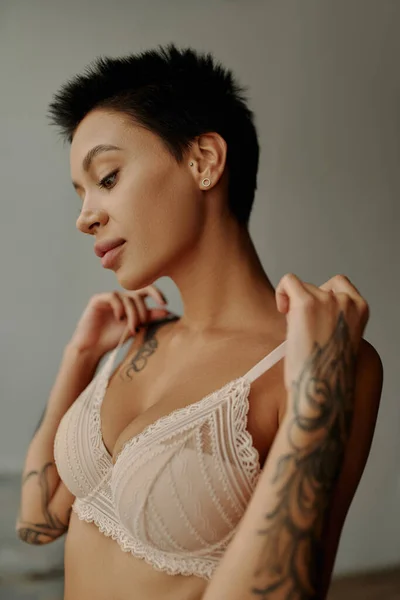 Sexy mujer tatuada tocando correas de sujetador en casa - foto de stock