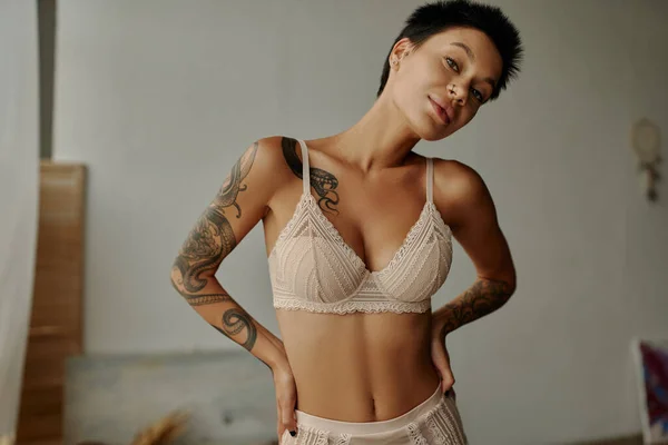 Young tattooed woman in beige lingerie looking at camera at home — Stock Photo