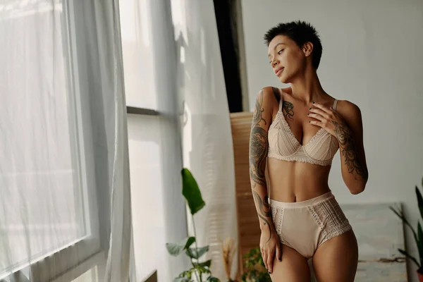 Jeune femme tatouée en lingerie dentelle regardant la fenêtre à la maison — Photo de stock