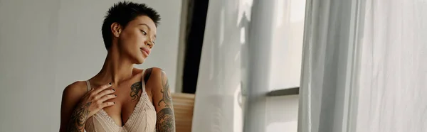 Sexy femme tatouée en soutien-gorge debout près de la fenêtre à la maison, bannière — Photo de stock