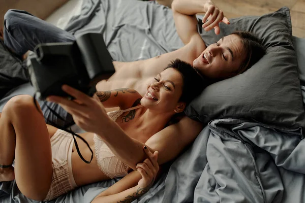 Top view of smiling woman in lingerie taking selfie with shirtless boyfriend on bed — Stock Photo