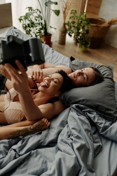 Sexy woman in bra taking selfie on vintage camera near young boyfriend on bed — Stock Photo
