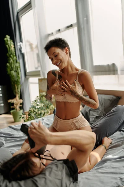 Мужчина без рубашки фотографирует сексуальную подружку на винтажную камеру в спальне — стоковое фото