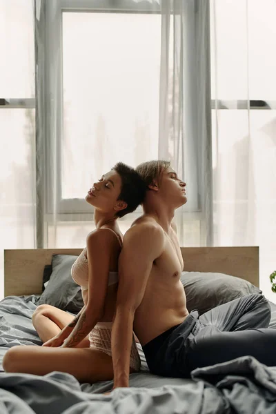 Homme torse nu en pyjama pantalon assis dos à dos avec petite amie sexy sur le lit — Photo de stock