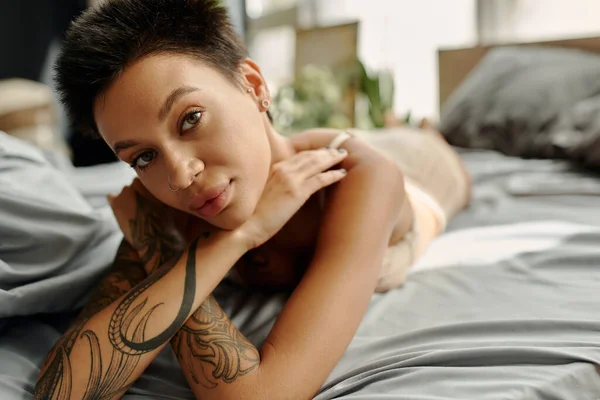 Молода татуйована жінка дивиться на камеру, лежачи на ліжку — стокове фото