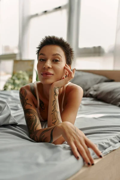Sexy mujer tatuada mirando a la cámara mientras está acostada en la cama por la mañana - foto de stock