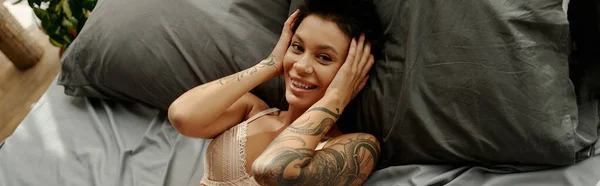 High angle view of cheerful tattooed woman in bra looking at camera on bed, banner — Stock Photo