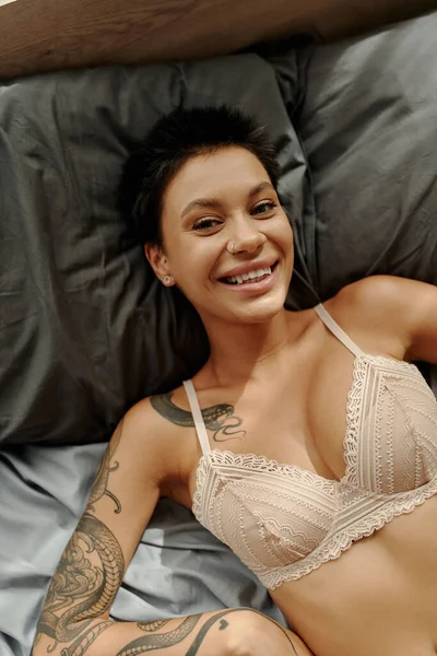 Top view of positive tattooed woman in lace bra looking at camera while lying on bed — Stock Photo