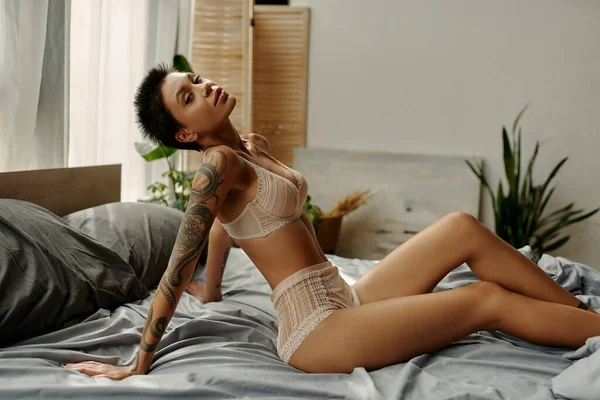 Татуированная женщина в бежевом белье смотрит в камеру на кровати — стоковое фото