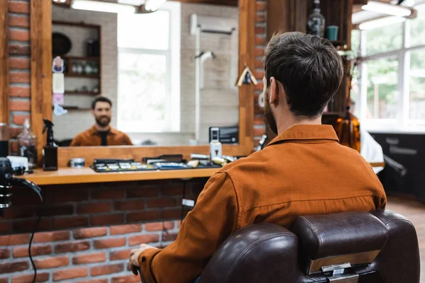 Vue arrière de l'homme brunette assis dans une chaise de coiffure près d'un miroir flou — Photo de stock