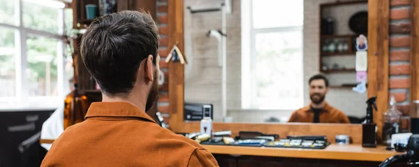 Rückansicht eines brünetten Mannes, der in einem Friseurladen in der Nähe eines verschwommenen Spiegels sitzt, Banner — Stockfoto