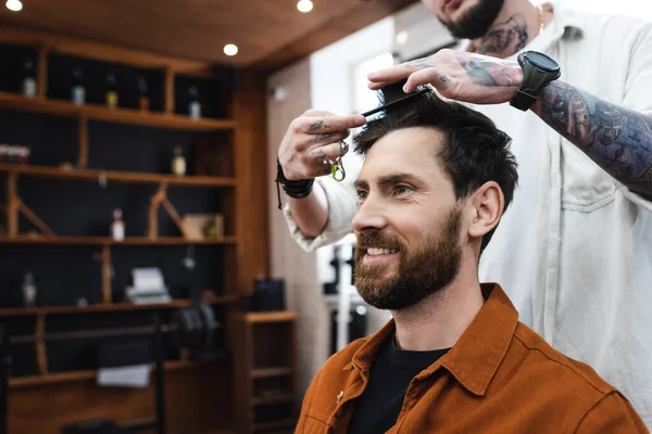 Cabeleireiro tatuado pentear o cabelo do homem barbudo sorridente na barbearia — Fotografia de Stock