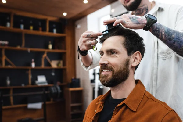 Tattooed hairdresser combing hair of smiling bearded man in barbershop — Stock Photo