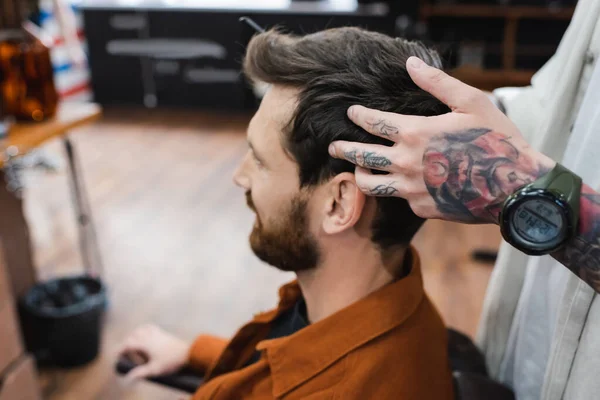 Peluquero tatuado en reloj de pulsera tocando el pelo de hombre morena en la barbería - foto de stock