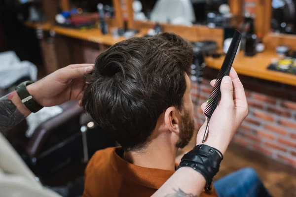 Tätowierter Friseur hält Kamm in der Nähe brünetten Mannes in Friseurladen — Stockfoto