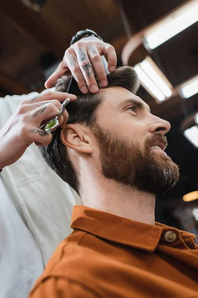 Low angle view of hairstylist cutting hair of bearded smiling man — Stock Photo