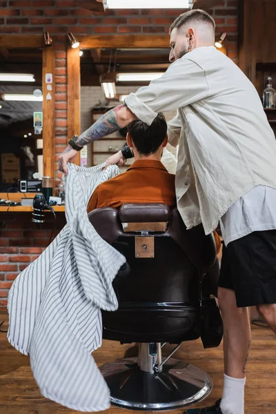 Tatuato barbiere mettere il mantello parrucchiere sul cliente seduto in poltrona nel salone di bellezza — Foto stock