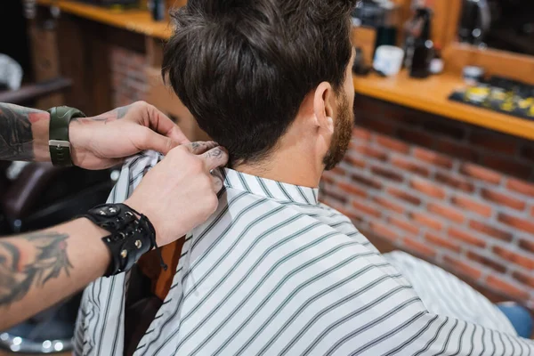Tattooed barber putting hairdressing cape on brunette man — Stock Photo