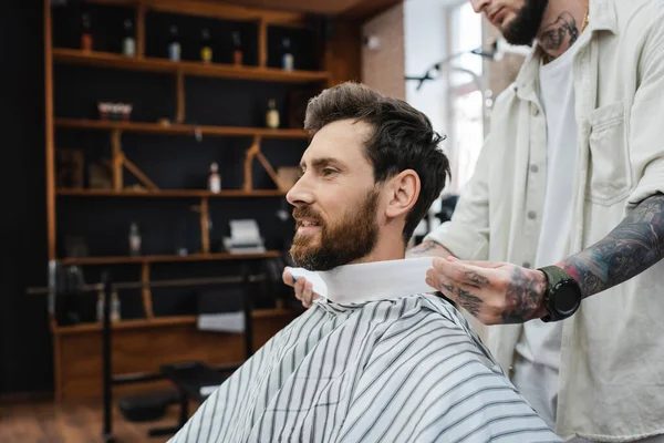 Tattooed barber wrapping neck of bearded man with hairdressing collar — Stock Photo