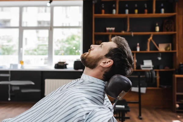 Vista laterale di uomo bruna barbuto seduto in parrucchiere mantello in barbiere — Foto stock
