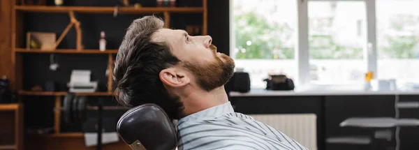 Side view of brunette bearded man in hairdressing cape in barbershop, banner — Stock Photo