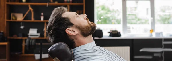 Vista lateral del hombre barbudo morena en la capa de peluquería en la barbería, pancarta — Stock Photo