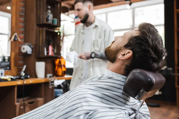 Selective focus of man in hairdressing cape sitting near blurred barber in beauty salon — Stock Photo