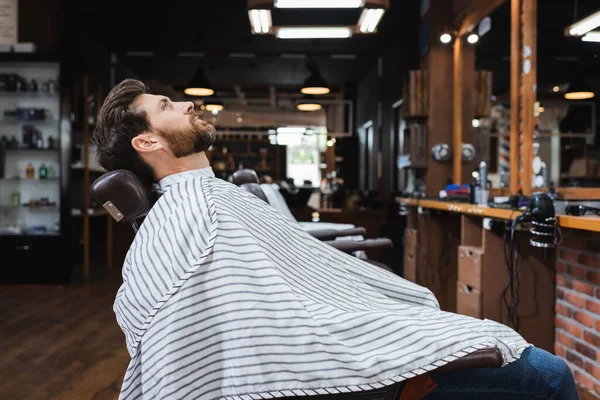 Vista lateral de morena barbudo hombre sentado en peluquería capa en salón de belleza - foto de stock