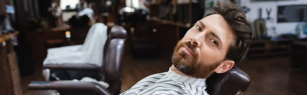 Hombre barbudo mirando a la cámara mientras está sentado en la peluquería borrosa, pancarta - foto de stock