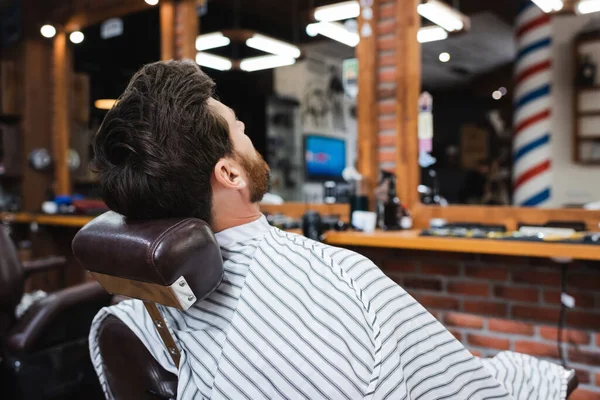 Homem morena na capa de cabeleireiro sentado perto de espelhos borrados na barbearia — Fotografia de Stock