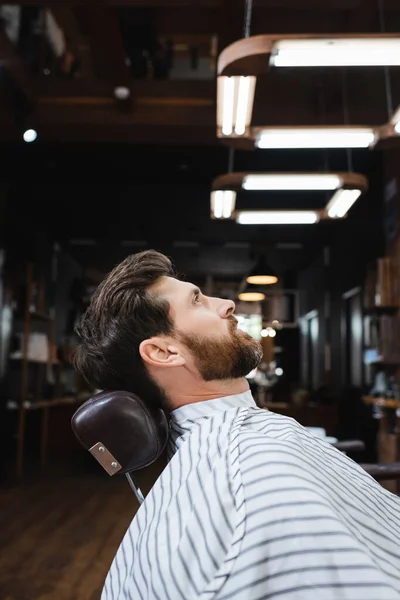 Vista lateral del hombre morena barbuda sentado en la capa de peluquería en la barbería - foto de stock