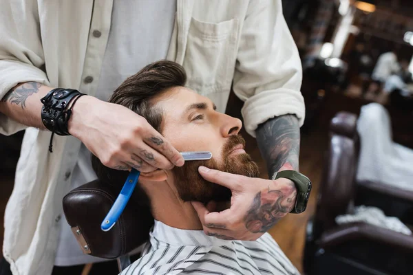 Tätowierter Friseur rasiert bärtigen Mann mit Rasiermesser — Stockfoto
