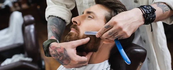 Tätowierter Friseur in Lederarmband rasiert Mann mit Rasiermesser, Banner — Stockfoto