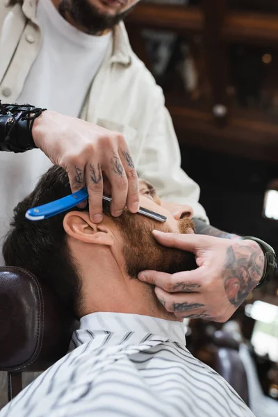 Tatuado cabeleireiro barbear bochecha do cliente com navalha reta — Stock Photo
