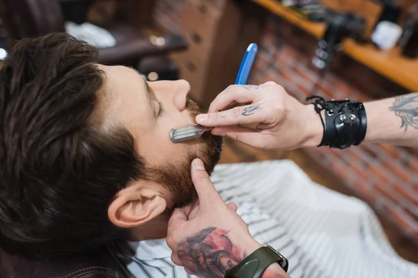Visão de alto ângulo de barbeiro tatuado barbeando homem barbudo com navalha reta — Stock Photo