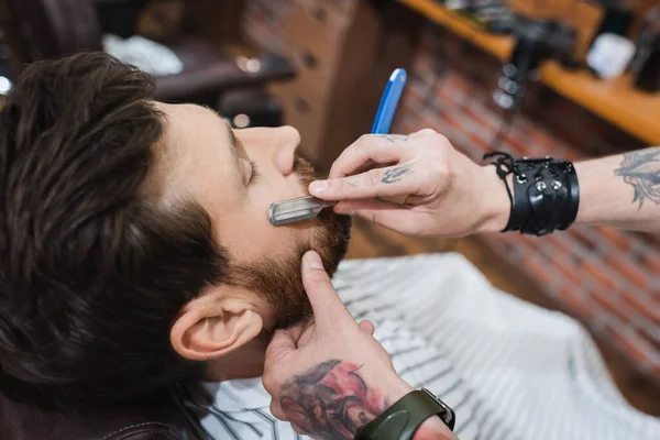 High angle view of tattooed barber shaving bearded man with straight razor — Stock Photo