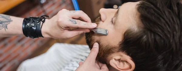 Hairdresser in leather bracelet shaving cheek of man with straight razor, banner — Stock Photo