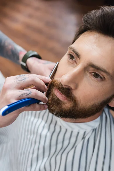 Vista de ángulo alto del hombre barbudo cerca de peluquero afeitándolo con navaja de afeitar recta - foto de stock