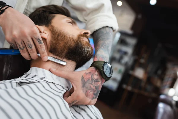 Low angle view of tattooed barber in wristwatch shaving neck of bearded man with straight razor — Stock Photo