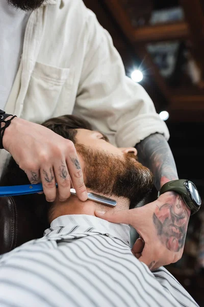 Tattooed hairdresser shaving neck of bearded man with straight razor — Stock Photo