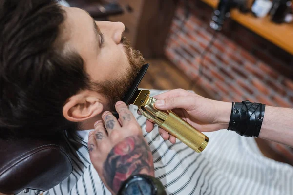 Vista de ángulo alto de peluquero tatuado barba de corte de hombre con cortador de pelo - foto de stock