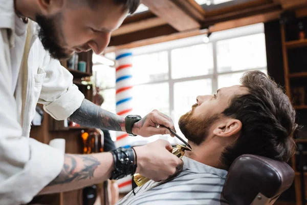 Morena barbudo hombre cerca de estilista trabajando con peine y cortador de pelo - foto de stock