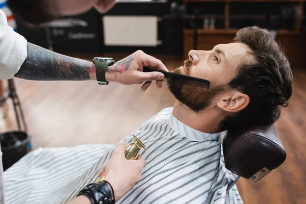 Barbeiro tatuado segurando cortador de cabelo e pentear a barba do homem na barbearia — Fotografia de Stock