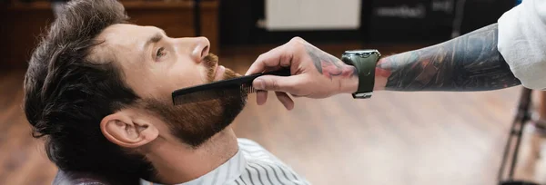Вид збоку татуйованого перукаря в наручнику розчісує бороду клієнта, банер — стокове фото